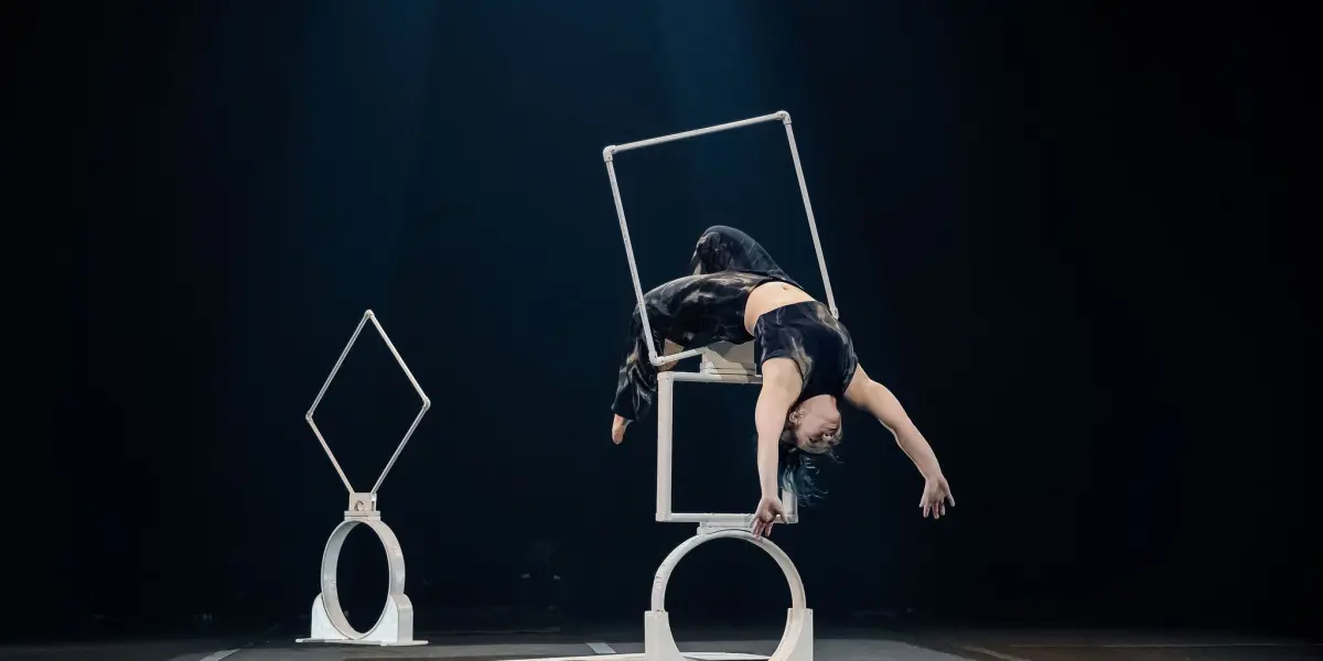 Un artiste vêtu de vêtements sombres, peut-être Haley Tenn, se tient en équilibre à l'envers sur une structure géométrique, le dos voûté et un bras tendu vers le bas, sur une scène faiblement éclairée, avec une autre structure en arrière-plan.