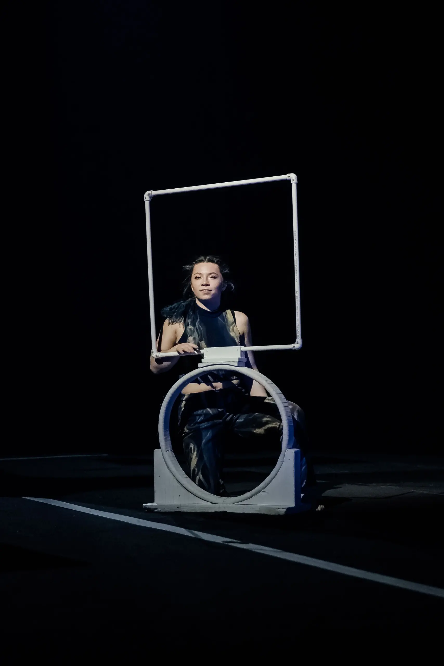 Un artiste, peut-être Haley Tenn, habillé de vêtements sombres, est assis à l'intérieur d'un cadre circulaire sur la scène, tenant un bâton surmonté d'un cadre carré. L'arrière-plan est sombre et l'éclairage dramatique se concentre sur l'artiste.