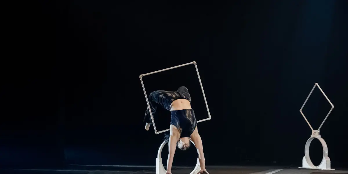 L'acrobate Haley Tenn se tient en équilibre sur ses mains au sommet d'un appareil circulaire blanc, les jambes repliées au-dessus d'un cadre carré, sur une scène faiblement éclairée, avec un autre appareil similaire en arrière-plan.