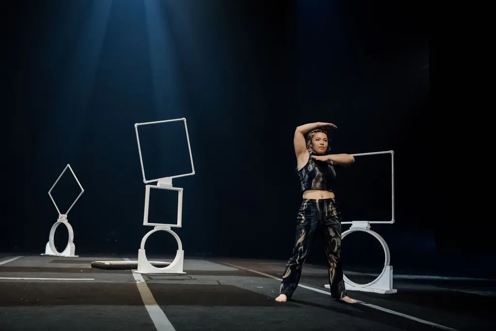 Une artiste de cirque, Haley Tenn, vêtue d'une tenue noire, prend la pose sur scène avec de grands accessoires géométriques, notamment des cercles et des carrés, sur un fond sombre et dramatique éclairé par des projecteurs.