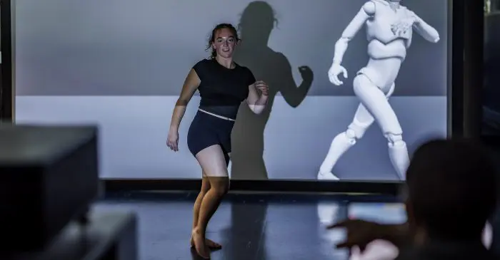 Une femme en tenue de sport danse devant un écran projeté avec une figure humanoïde 3D en mouvement à HUPR reconnu comme centre d'accès à la technologie, son ombre est projetée sur le mur tandis qu'une personne au premier plan fait un geste vers elle.