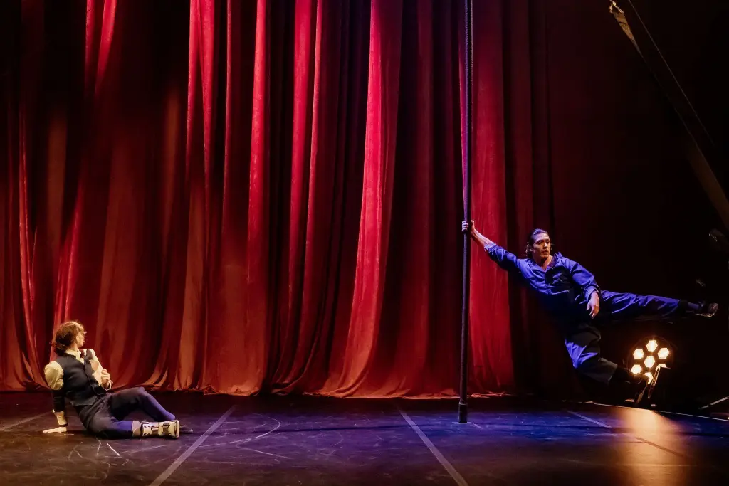 Deux artistes vêtus de tenues sombres se trouvent sur une scène entourée de rideaux rouges dramatiques. Yan Rocha Szuecs est allongée sur le sol, regardant l'autre s'accrocher à un poteau vertical, suspendant son corps à l'horizontale dans les airs. Les lumières de la scène brillent derrière eux.