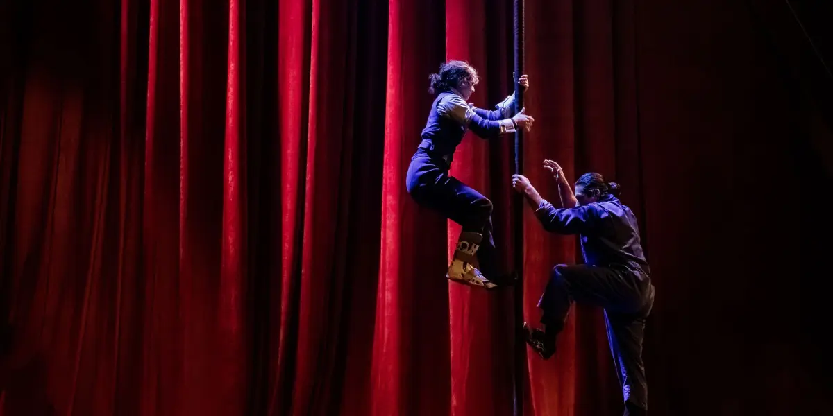 Deux artistes, dont Yan Rocha Szuecs, vêtus de tenues sombres, grimpent sur une corde verticale devant de grands rideaux de scène rouges et dramatiques, sous un éclairage théâtral.