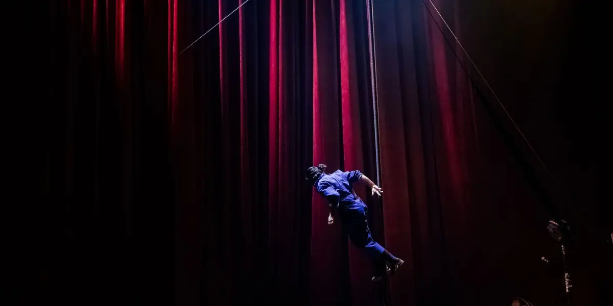 Yan Rocha Szuecs, vêtue de bleu, se tient en équilibre horizontal sur un poteau vertical sur la scène, encadrée par des rideaux de velours rouge dramatiques et des lumières de scène vives.