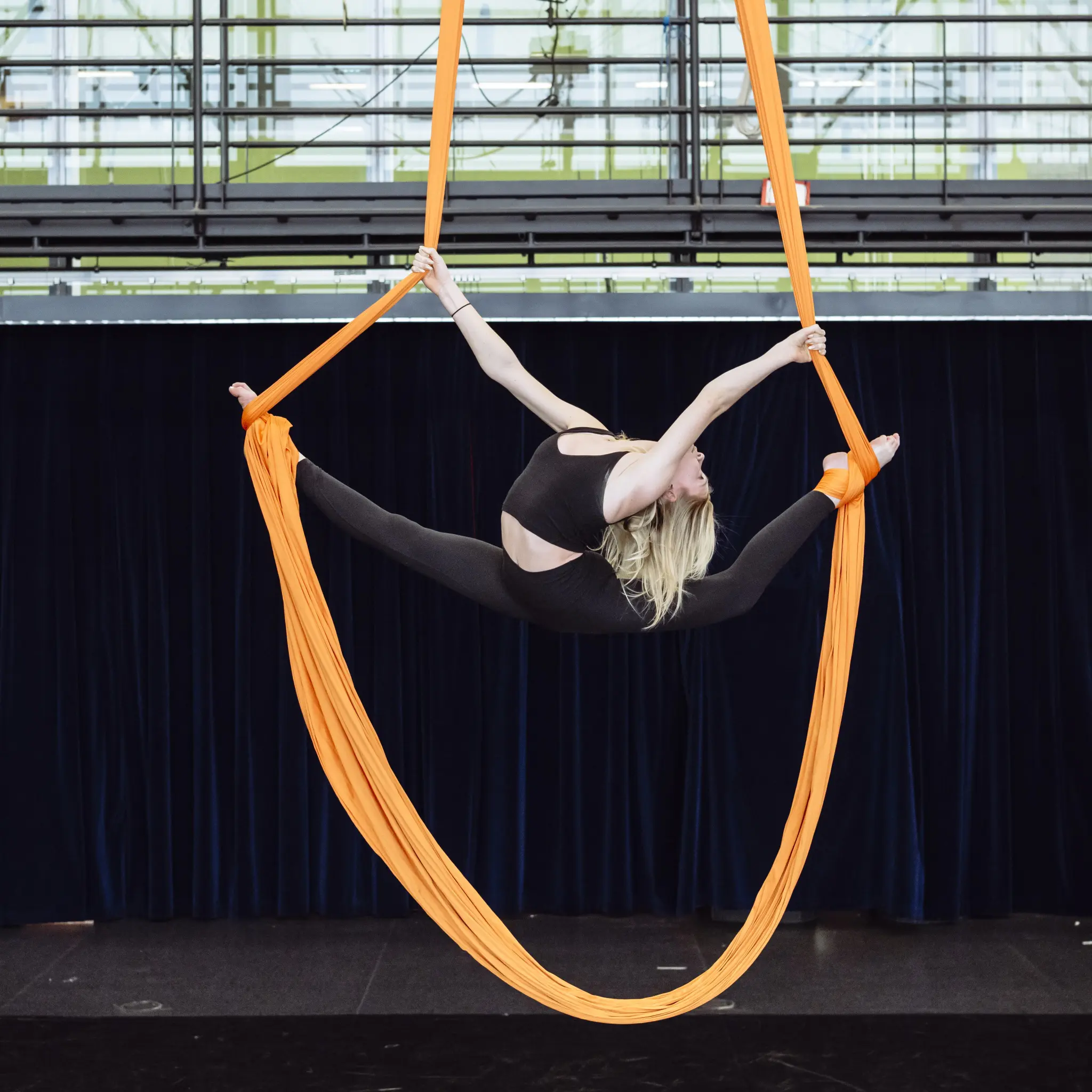Lors des cours récréatifs de l'ENC, une femme effectue un fractionnement aérien sur des soies orange, suspendue dans les airs, bras et jambes tendus. Elle porte une tenue noire, avec un rideau bleu foncé en toile de fond.