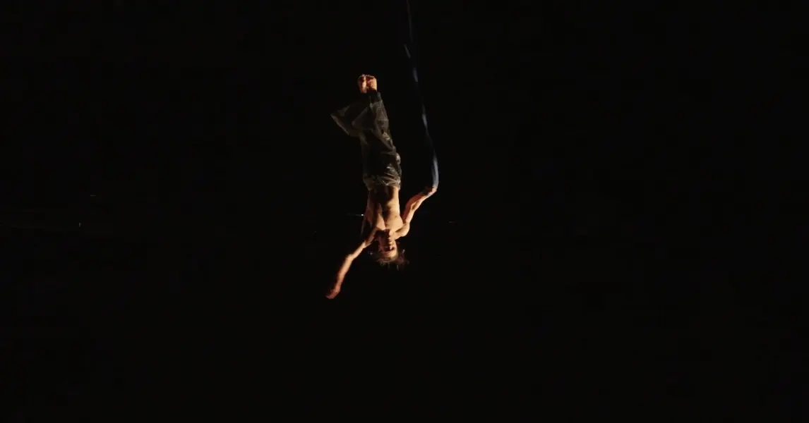 Une personne exécute un numéro de soie aérienne, suspendue à l'envers dans les airs sur un fond sombre, les bras étendus et les jambes enveloppées de tissu, rappelant les performances de Yan Rocha Szuecs.
