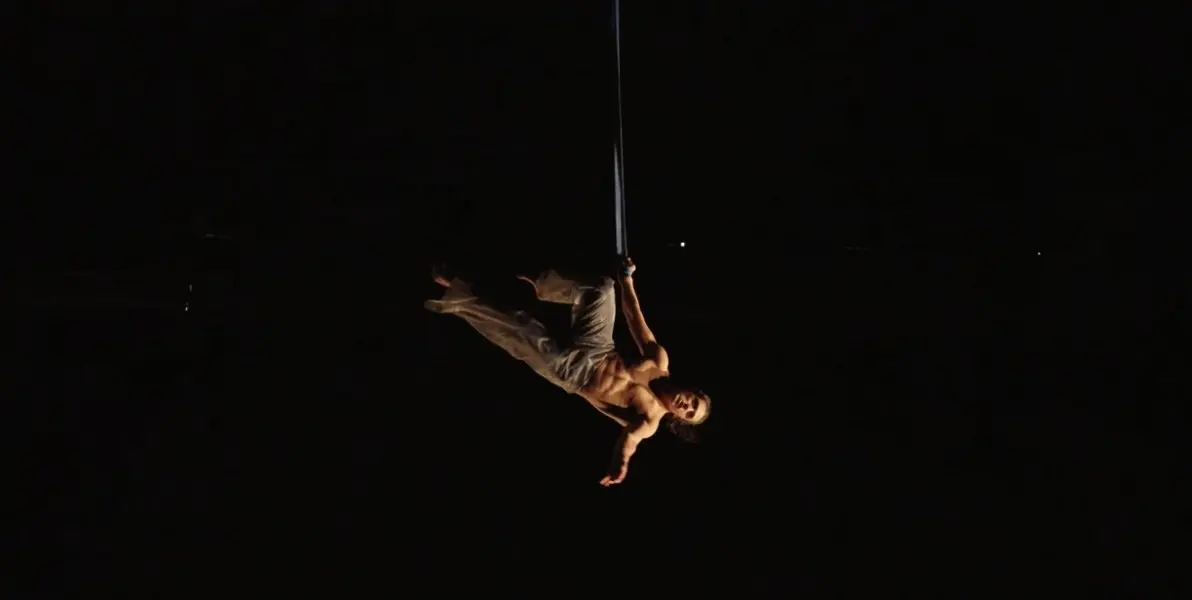 Yan Rocha Szuecs exécute un numéro aérien, suspendue à un tissu ou à une corde sur un fond sombre, vêtue d'un pantalon ample de couleur claire et sans chemise, un bras tendu vers le bas.