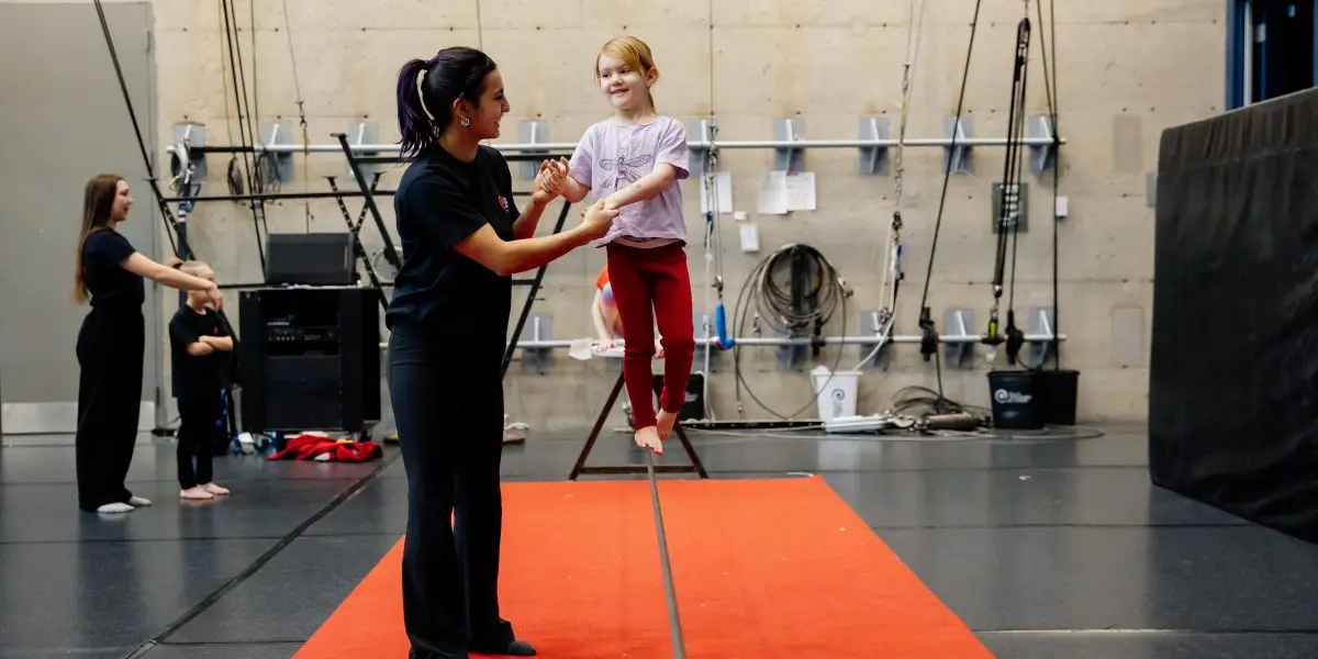 Une jeune fille se tient en équilibre sur une slackline avec le soutien d'un moniteur. Ils sont à l'intérieur sur un tapis rouge, et un autre instructeur et un autre enfant sont visibles à l'arrière-plan. Le décor semble être celui d'un gymnase ou d'un centre d'entraînement.