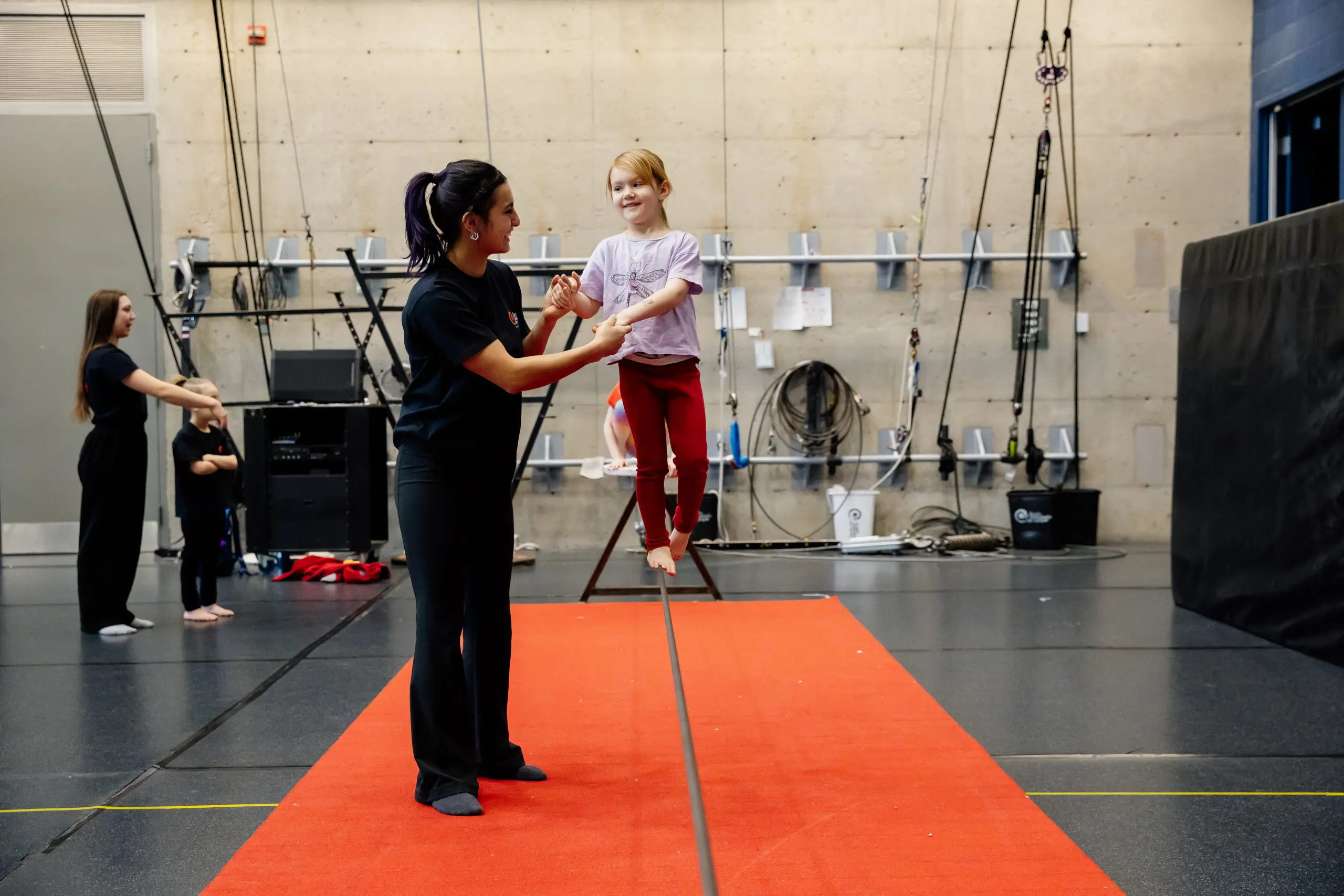 Une jeune fille se tient en équilibre sur une slackline avec le soutien d'un moniteur. Ils sont à l'intérieur sur un tapis rouge, et un autre instructeur et un autre enfant sont visibles à l'arrière-plan. Le décor semble être celui d'un gymnase ou d'un centre d'entraînement.