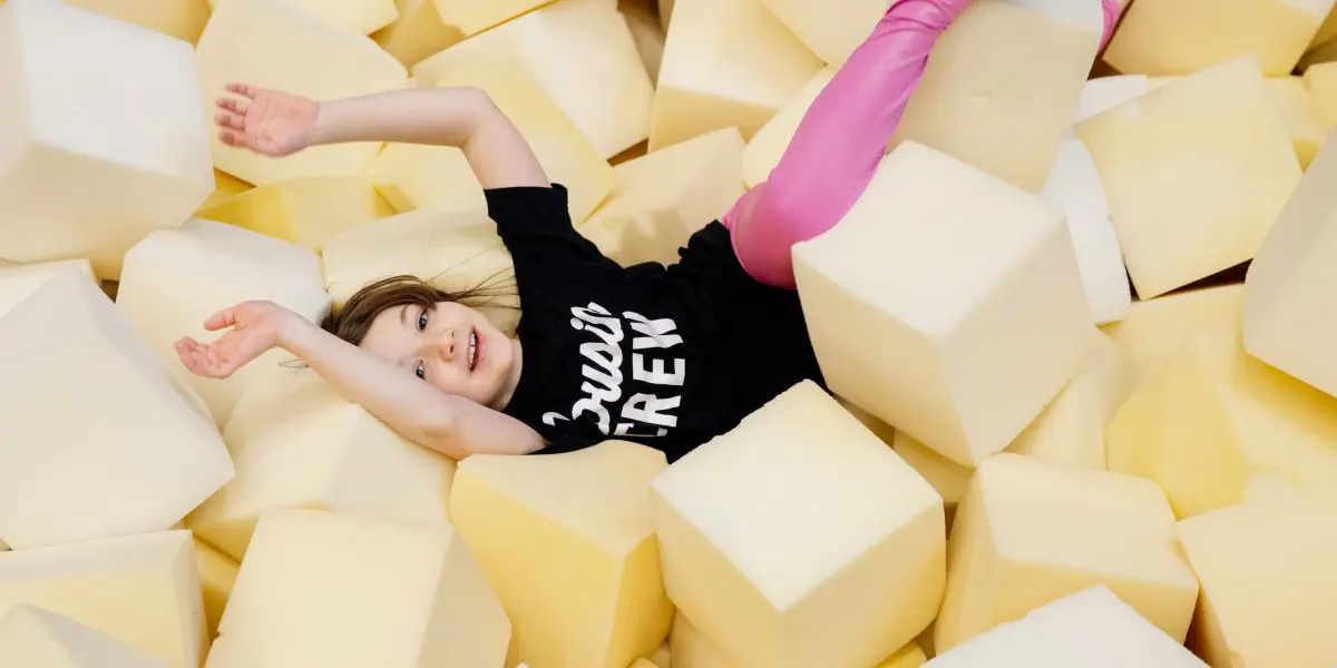 Un jeune enfant vêtu d'une chemise noire et d'un legging rose sourit et est allongé sur le dos dans une fosse remplie de gros cubes en mousse souple, les bras et les jambes tendus de manière ludique.