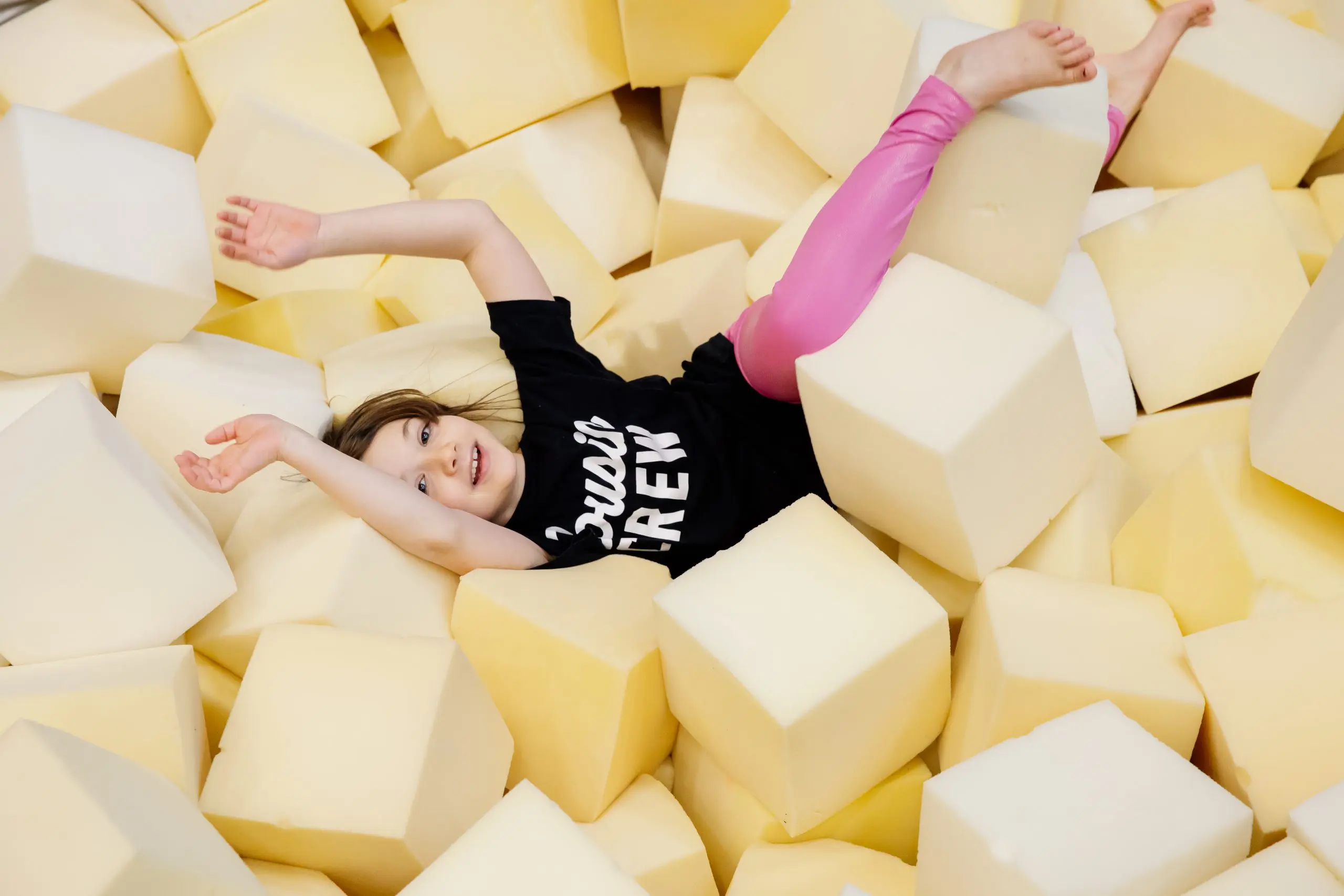 Un jeune enfant vêtu d'une chemise noire et d'un legging rose sourit et est allongé sur le dos dans une fosse remplie de gros cubes en mousse souple, les bras et les jambes tendus de manière ludique.