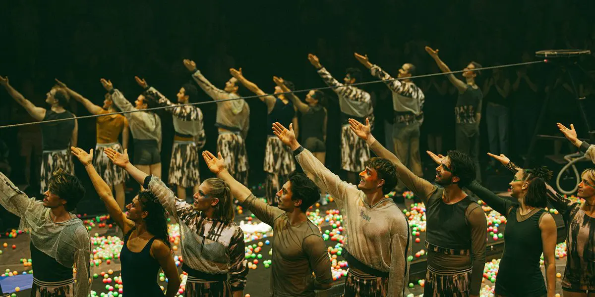Un groupe d'artistes se tient en rangs sur une scène couverte de boules colorées, étendant leurs bras droits vers le haut à l'unisson lors de la soirée-bénéfice de la Fondation de l'ENC, sous le regard d'un public en arrière-plan faiblement éclairé.