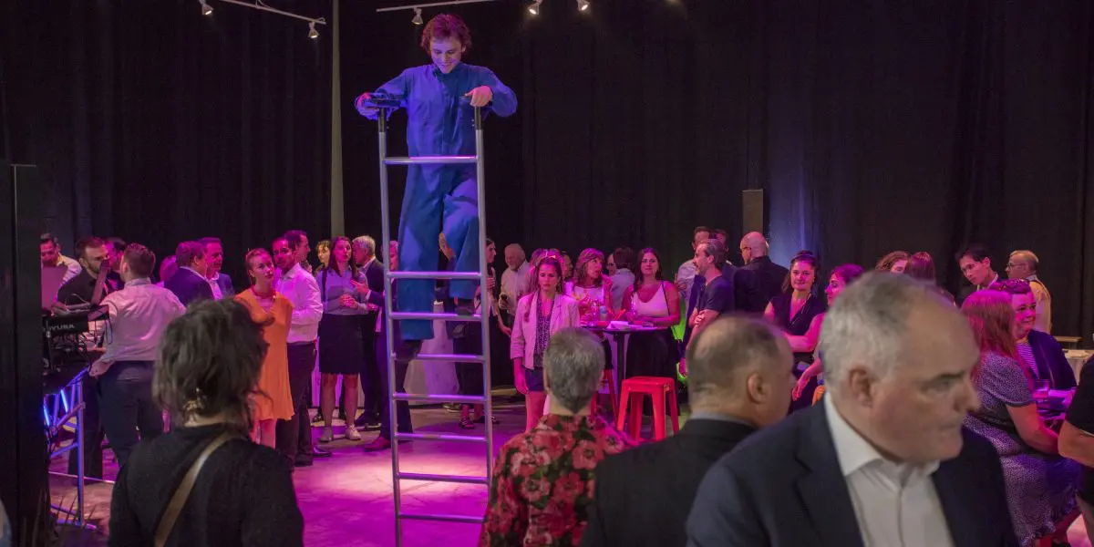 Un artiste en bleu de travail se tient sur une grande échelle au milieu d'une salle bondée lors de la soirée-bénéfice de la Fondation de l'ENC. Des éclairages colorés projettent des teintes violettes et roses sur la scène animée.