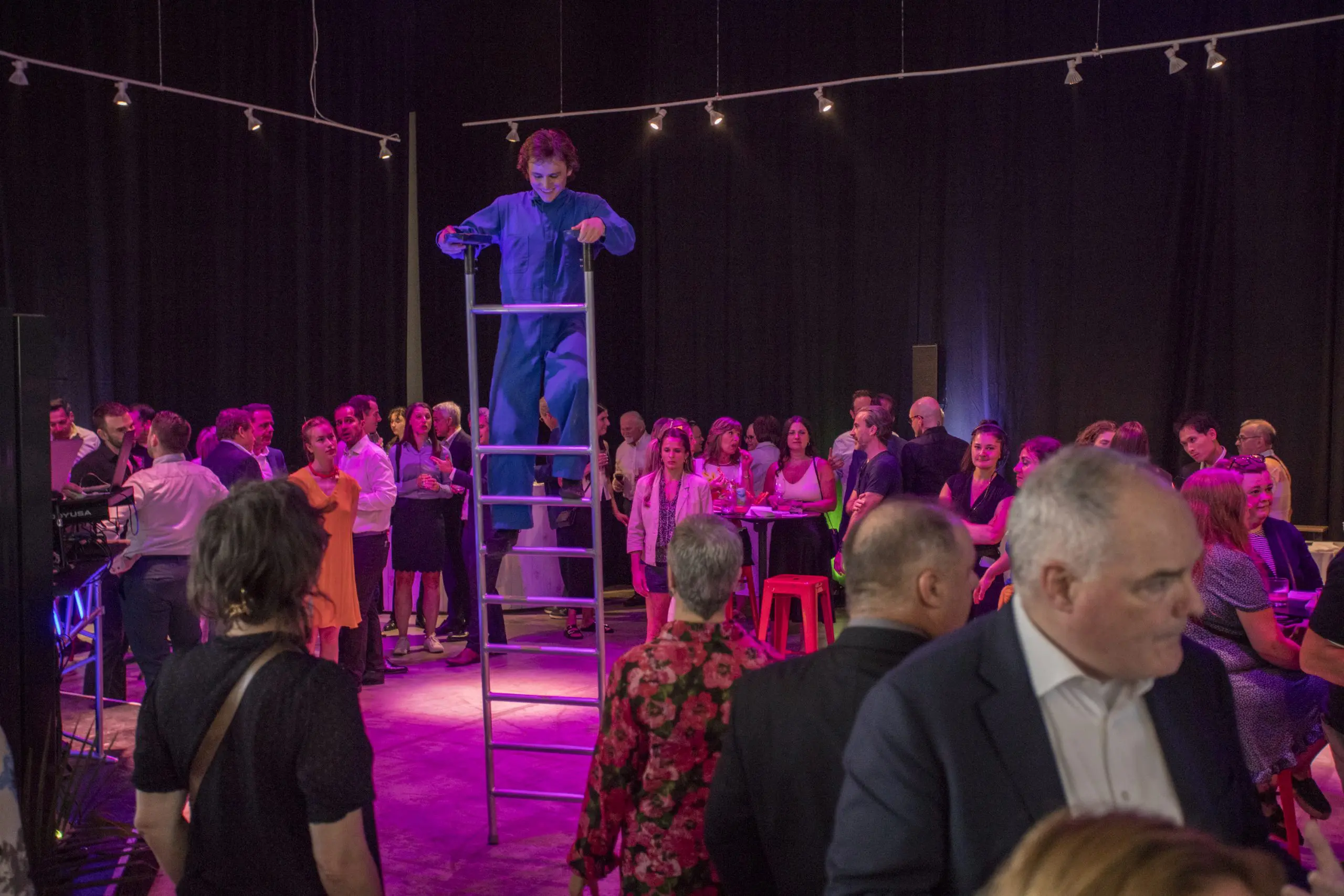 Un artiste en bleu de travail se tient sur une grande échelle au milieu d'une salle bondée lors de la soirée-bénéfice de la Fondation de l'ENC. Des éclairages colorés projettent des teintes violettes et roses sur la scène animée.