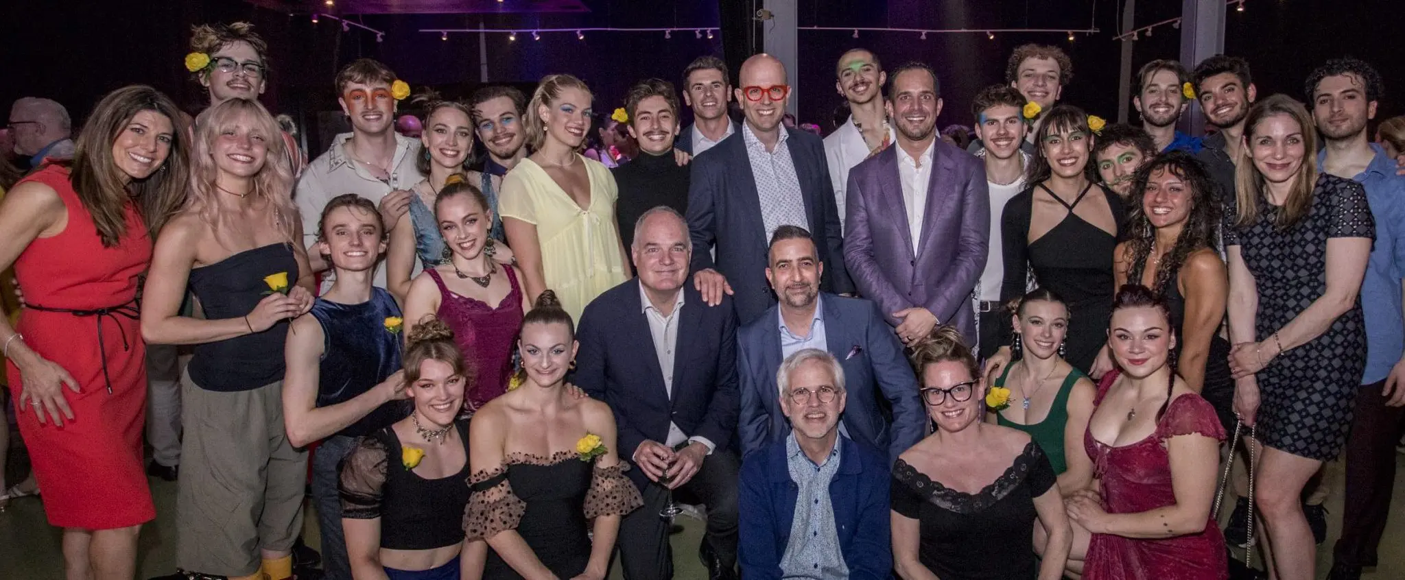 Un grand groupe de personnes vêtues de tenues de fête colorées et élégantes posent ensemble, souriant à l'appareil photo lors de la soirée-bénéfice de la Fondation de l'ENC. Certains portent des lunettes brillantes et des accessoires floraux, avec un éclairage festif en arrière-plan.