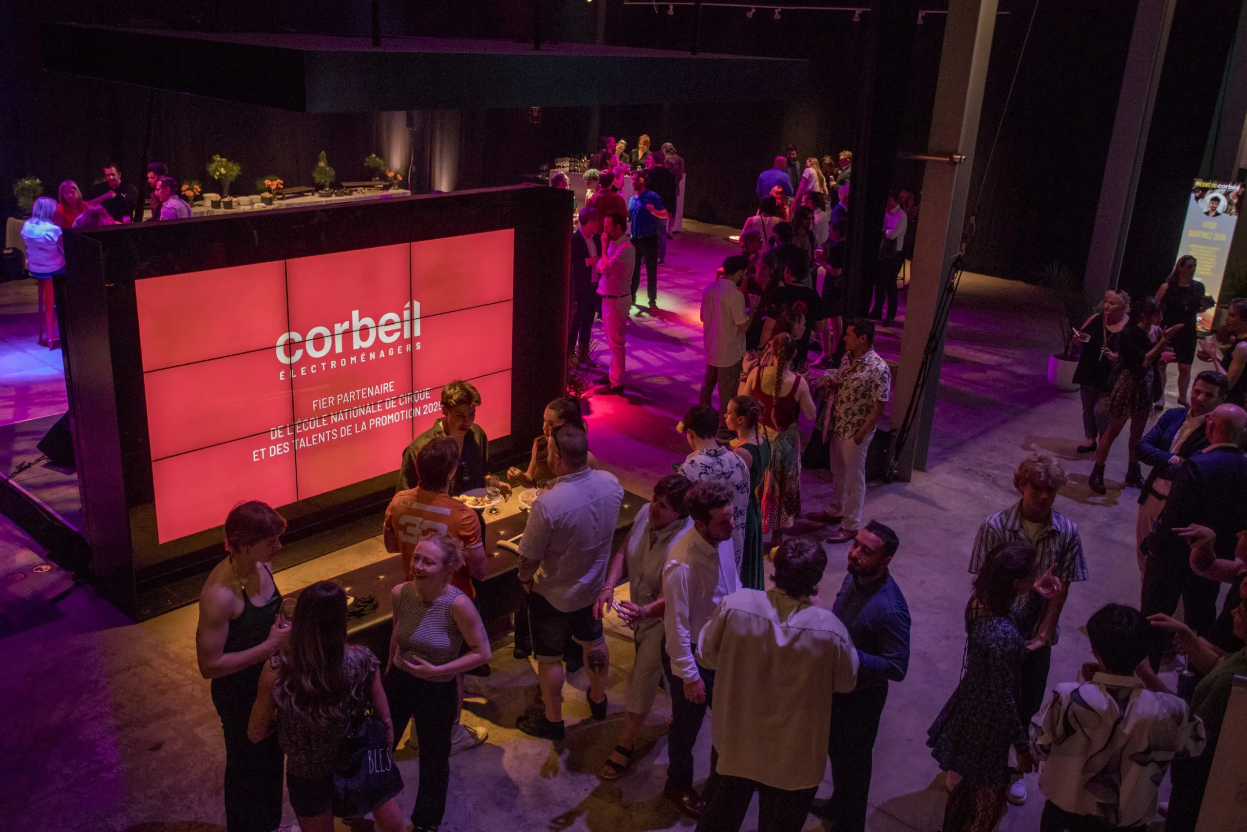 Des personnes se réunissent et socialisent lors d'une soirée-bénéfice en intérieur de la Fondation de l'ENC sous un éclairage violet. Un grand écran numérique rouge affiche des corbeils électroménagers et des textes en français tandis que les invités se mélangent, debout ou assis à des tables.