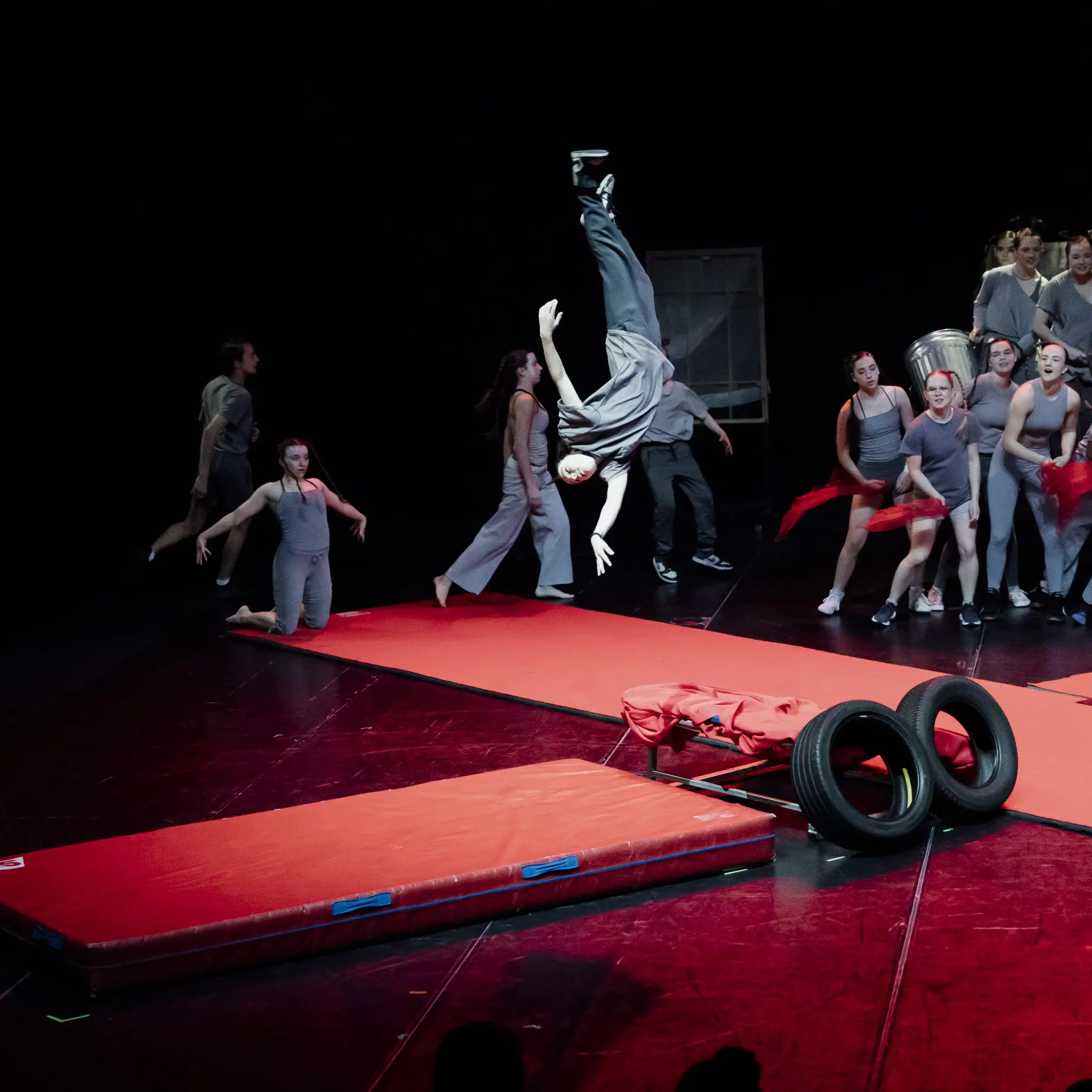 Un groupe d'artistes du programme préparatoire des arts du cirque monte sur scène - certains en costumes gris, l'un d'entre eux faisant un flip à mi-hauteur sur un tapis rouge. D'autres observent, tenant des tissus rouges parmi des accessoires tels que des pneus et des couvercles de poubelle argentés.