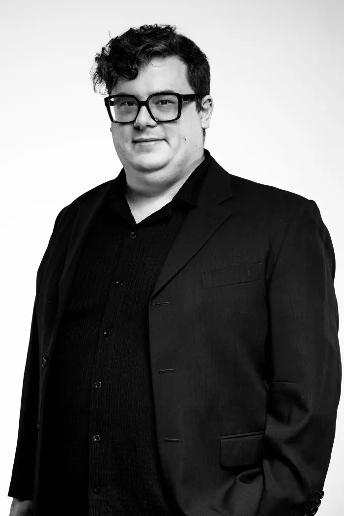 Un homme aux cheveux courts et bouclés et aux lunettes, identifié comme Olivier Leblanc, porte une veste de costume et une chemise sombres, debout sur un fond clair uni. L'image est en noir et blanc.