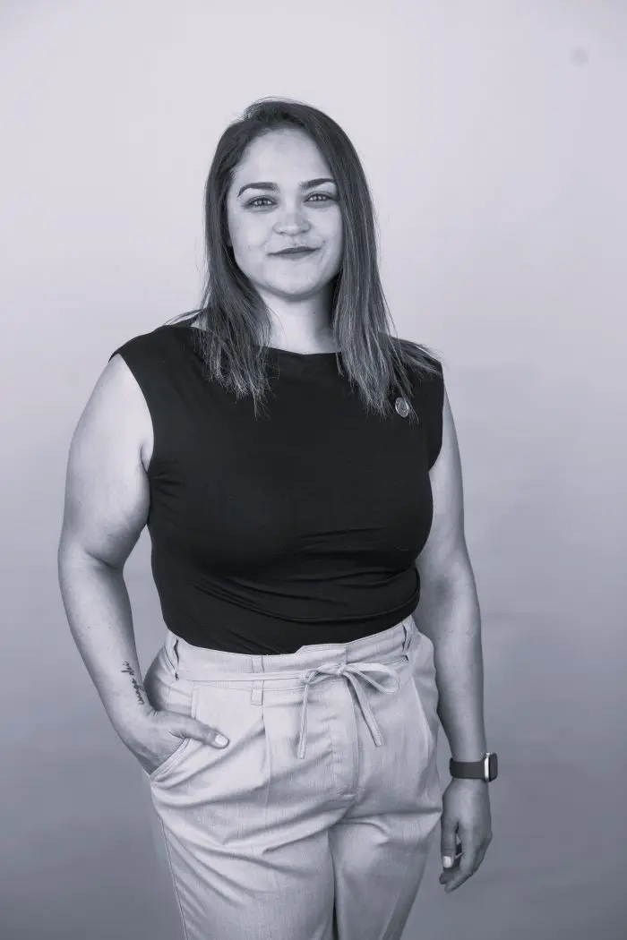 Mélanie-Beby Robert, aux cheveux raides tombant jusqu'aux épaules, porte un haut noir sans manches et un pantalon clair à taille haute ; elle se tient debout, une main dans la poche, et sourit à l'appareil photo devant un fond uni. La photo est en noir et blanc.