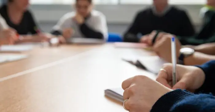 Gros plan d'une personne écrivant dans un cahier à une table de réunion, avec plusieurs personnes assises et discutant en arrière-plan - idéal pour illustrer un appel de candidatures - Comité d'éthique de la recherche en cours.
