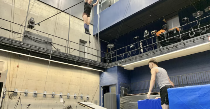 Une personne effectue un numéro aérien à l'aide d'un trapèze dans une salle de sport avec des tapis bleus en dessous, tandis qu'une autre personne vêtue d'un débardeur observe depuis le sol. L'espace est doté de hauts plafonds, de projecteurs et de cordes d'escalade sur le mur.