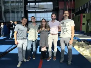 Cinq adultes se tiennent debout sur un tapis de gymnastique bleu dans un gymnase intérieur bien éclairé, posant pour une photo de groupe pendant la Semaine québécoise des directions d'établissements scolaires. Les blocs de mousse et l'équipement de gymnastique sont visibles grâce à la lumière du soleil qui pénètre par de grandes fenêtres.