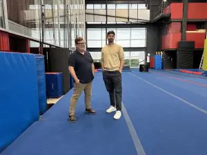 Deux personnes se tiennent sur un tapis de gymnastique bleu à l'intérieur d'un gymnase spacieux aux murs rouges et noirs, avec des équipements de gymnastique et de grandes fenêtres en arrière-plan, et sourient à la caméra pendant la Semaine québécoise des directions d'établissements scolaires.