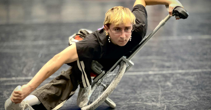 Une personne aux cheveux blonds courts et aux lunettes effectue une manœuvre athlétique au sol à l'aide d'un fauteuil roulant, en se penchant intensément d'un côté - montrant l'esprit des nouveaux cours récréatifs à l'École nationale de cirque !.