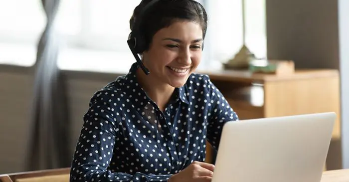 Une femme portant un casque et une chemise à pois bleu marine sourit alors qu'elle est assise à un bureau et qu'elle utilise un ordinateur portable. Elle semble être en appel vidéo ou travailler à distance dans une pièce lumineuse.