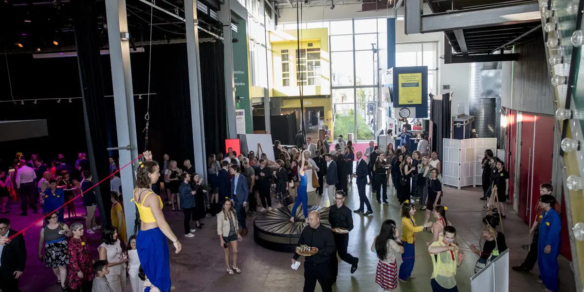 Une soirée-bénéfice intérieure animée de la Fondation de l'ENC avec des gens qui socialisent ; certains regardent des artistes sur une slackline et un cerceau aérien. Le lieu est doté de hauts plafonds, de grandes fenêtres, d'un décor industriel, de murs colorés et d'éclairages dispersés.