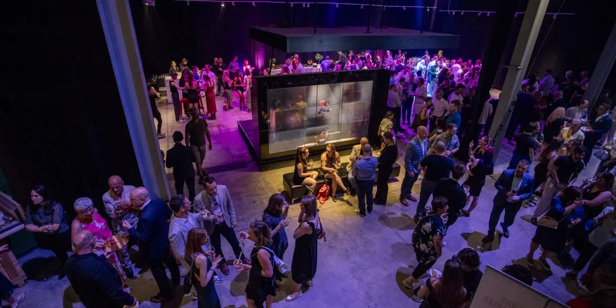 Une foule nombreuse socialise et se mêle à un événement en intérieur avec un éclairage tamisé et des projecteurs colorés violets et jaunes, rassemblés autour d'un affichage central illuminé pendant la soirée-bénéfice de la Fondation de l'ENC.
