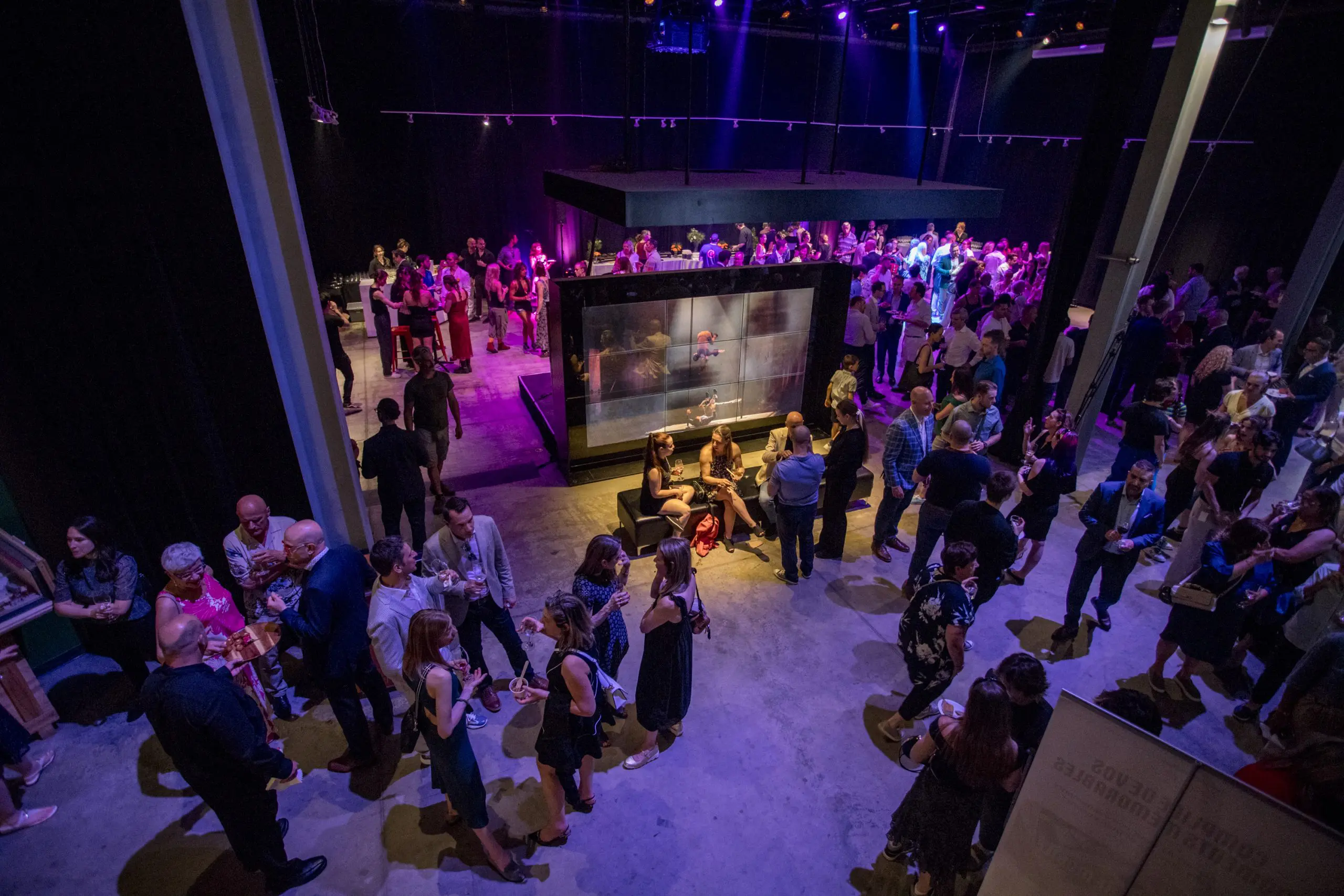 Une foule nombreuse socialise et se mêle à un événement en intérieur avec un éclairage tamisé et des projecteurs colorés violets et jaunes, rassemblés autour d'un affichage central illuminé pendant la soirée-bénéfice de la Fondation de l'ENC.