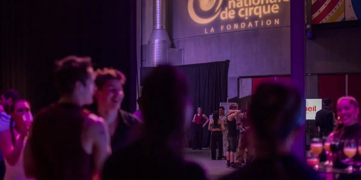 Les gens se rassemblent et socialisent lors d'une soirée-bénéfice de la Fondation de l'ENC sous un éclairage violet, avec le logo de l'École nationale de cirque LA FONDATION projeté sur le mur à l'arrière-plan.