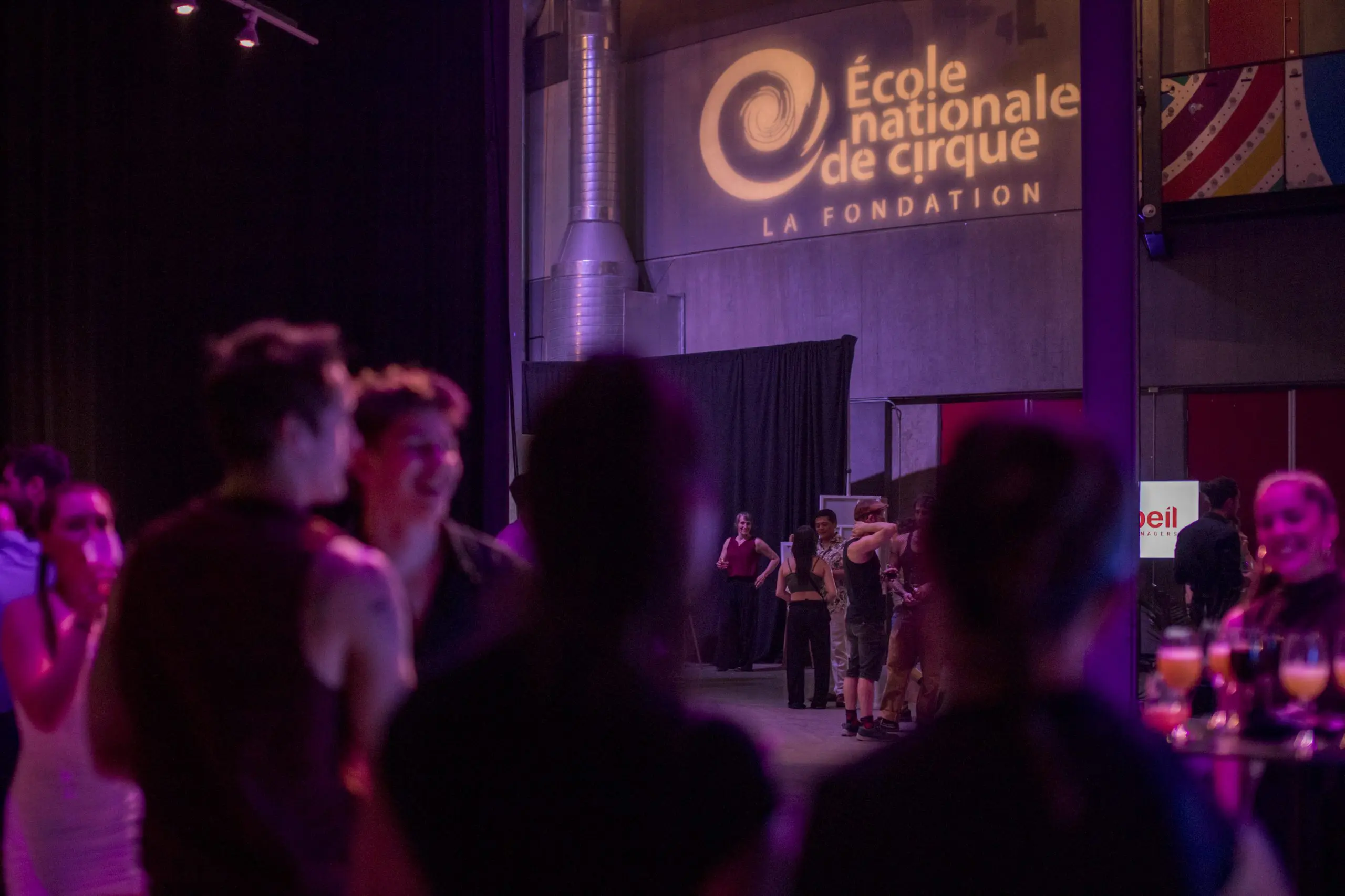 Les gens se rassemblent et socialisent lors d'une soirée-bénéfice de la Fondation de l'ENC sous un éclairage violet, avec le logo de l'École nationale de cirque LA FONDATION projeté sur le mur à l'arrière-plan.