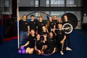 Un groupe de douze personnes vêtues de chemises noires assorties pose à l'intérieur avec des accessoires de cirque, notamment un monocycle, un cerceau, des massues de jonglage et une roue Cyr, tout en souriant à l'appareil photo dans un cadre semblable à celui d'un gymnase.