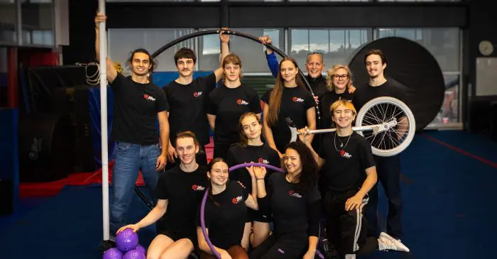 Un groupe de douze personnes vêtues de chemises noires assorties pose à l'intérieur avec des accessoires de cirque, notamment un monocycle, un cerceau, des massues de jonglage et une roue Cyr, tout en souriant à l'appareil photo dans un cadre semblable à celui d'un gymnase.