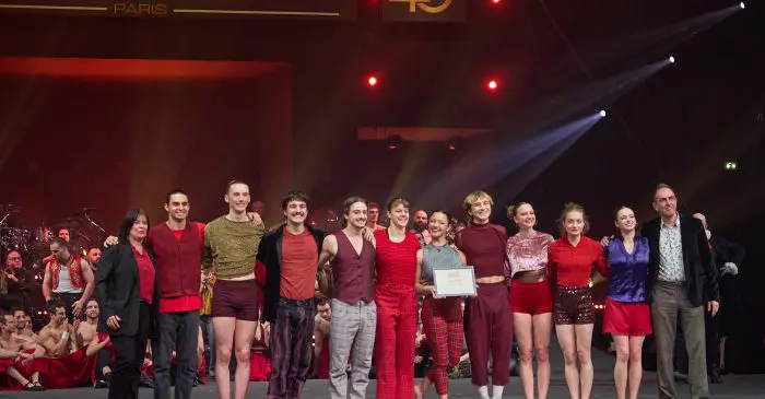Un groupe d'artistes en costumes rouge et or se tient sur scène, souriant et posant avec un certificat de récompense lors du 45e Festival Mondial du Cirque de Demain à Paris.