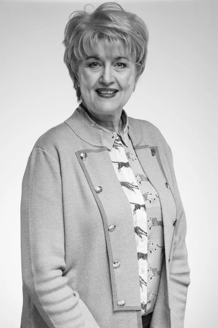 Une femme souriante d'un certain âge, aux cheveux courts et clairs, porte un chemisier à motifs et un gilet boutonné. Elle se tient debout sur un fond uni, regardant directement l'appareil photo. La photo est en noir et blanc.