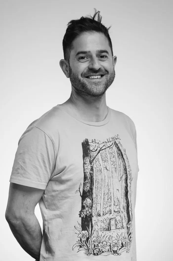 Un homme souriant aux cheveux bruns courts et à la barbe se tient debout sur un fond uni. Il porte un tee-shirt de couleur claire avec un arbre et une porte illustrés sur le devant. La photo est en noir et blanc.