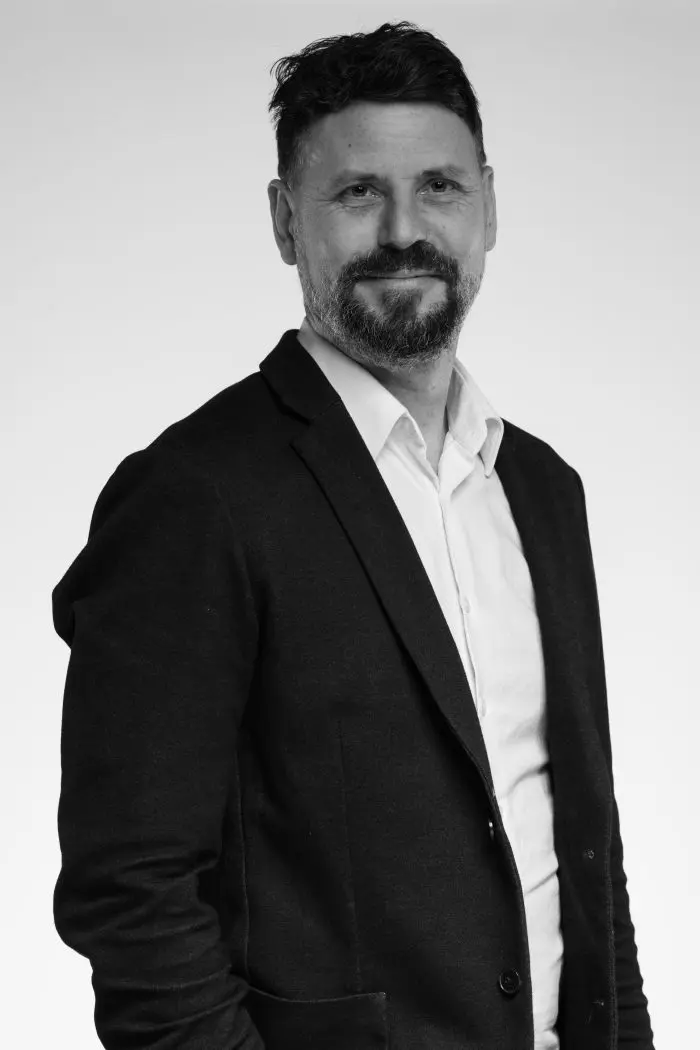 Photo en noir et blanc d'un homme aux cheveux et à la barbe courts, portant une veste de costume sur une chemise boutonnée, debout et souriant légèrement sur un fond uni.