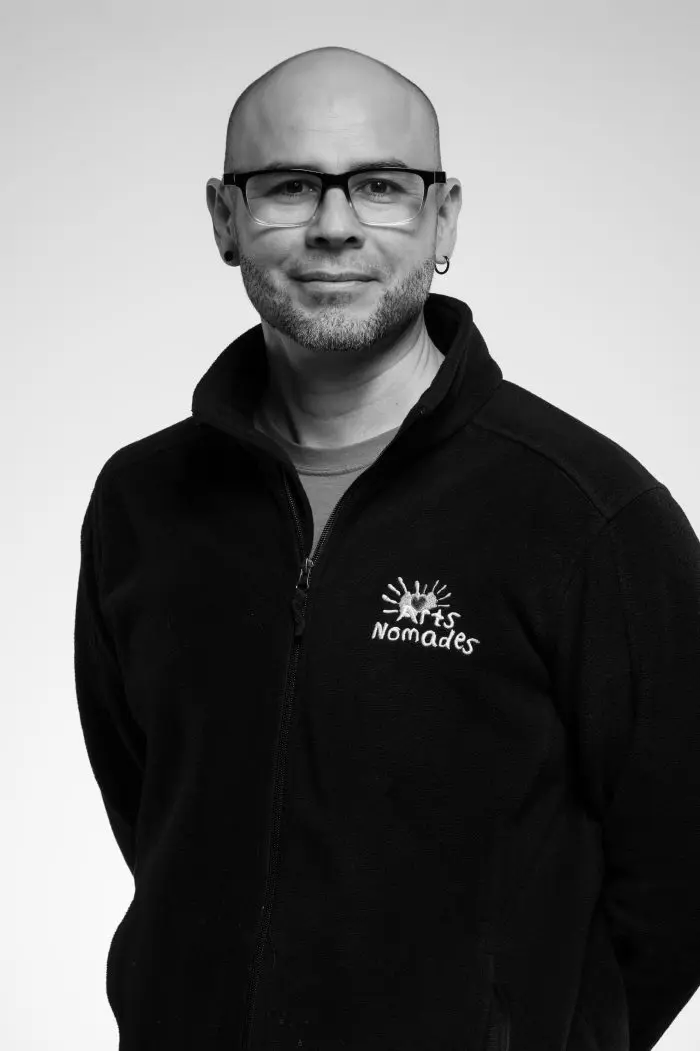 Un homme avec des lunettes et une courte barbe, portant une veste zippée noire avec un logo Arts Nomades, se tient debout sur un fond clair et uni. L'image est en noir et blanc.