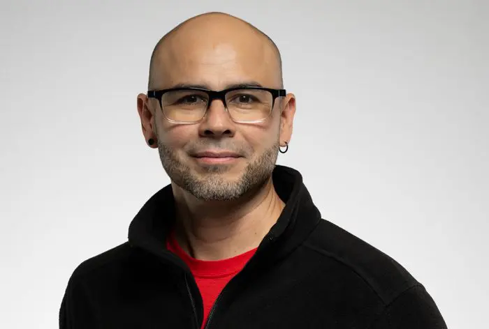 Un homme au crâne dégarni, portant des lunettes, une courte barbe et des boucles d'oreilles, est vêtu d'une veste noire sur une chemise rouge. Il sourit légèrement et pose sur un fond gris clair uni.