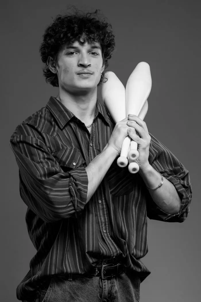 Photo en noir et blanc d'une jeune personne aux cheveux bouclés, portant une chemise rayée, tenant trois massues de jonglage sur son épaule et regardant l'appareil photo avec assurance.
