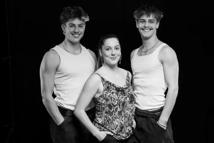 Trois jeunes adultes posent ensemble sur un fond noir. Deux hommes vêtus de débardeurs blancs se tiennent de part et d'autre d'une femme vêtue d'un haut sans manches à motifs. Tous sourient et ont les mains dans les poches.