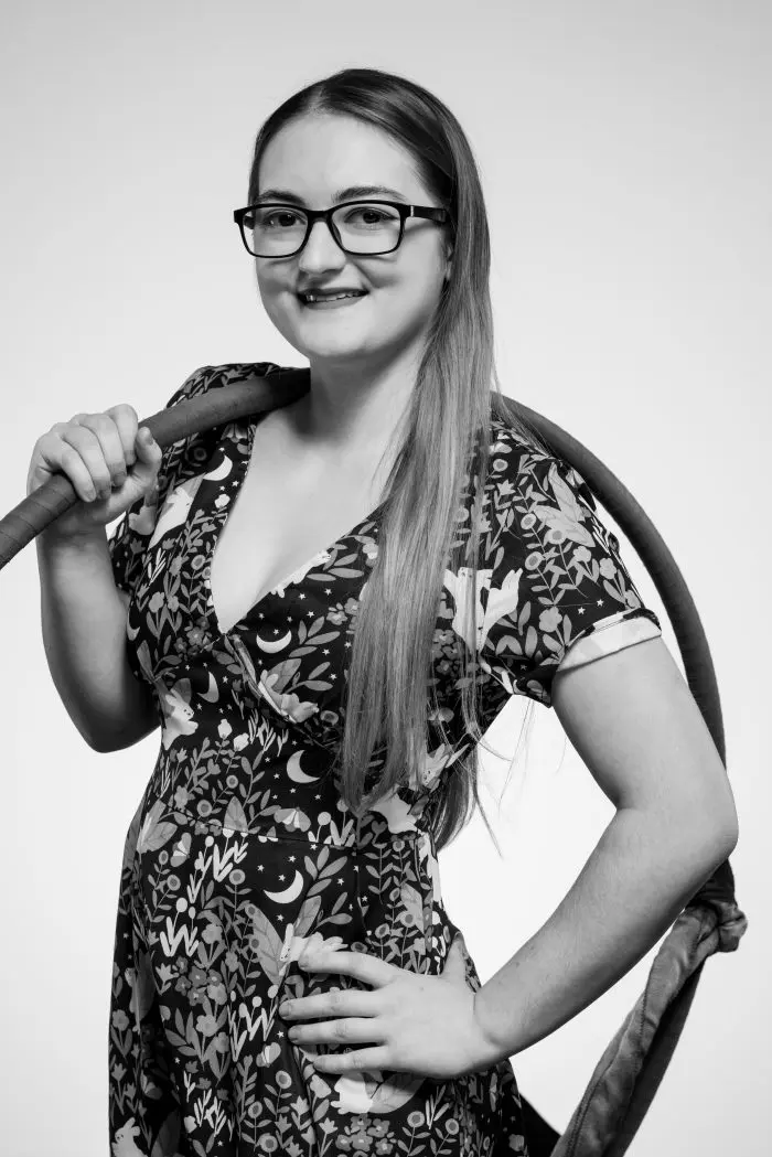 Une femme aux longs cheveux raides et aux lunettes sourit avec assurance, tenant un hula hoop sur son épaule. Elle porte une robe à motifs et à manches courtes. La photo est en noir et blanc sur un fond uni.