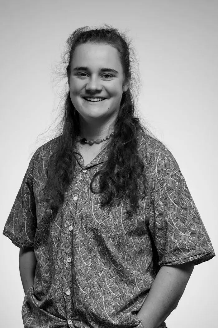 Portrait en noir et blanc d'une personne aux cheveux longs, souriante, portant une chemise à boutons à motifs, les mains dans les poches, debout sur un fond uni.