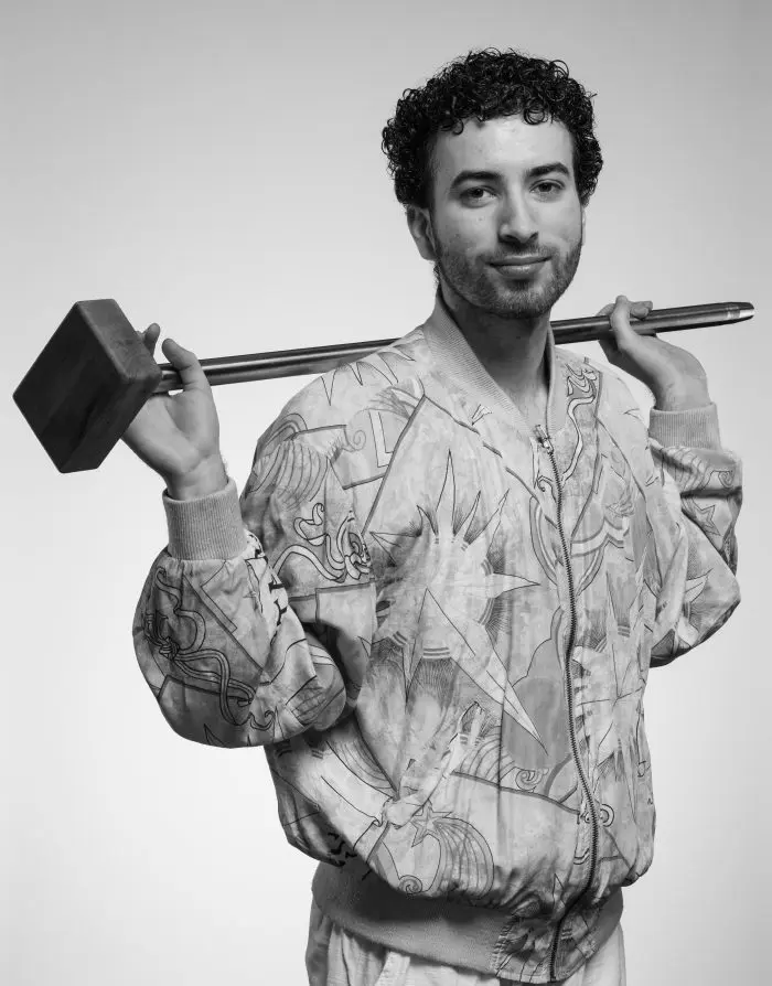Un homme aux cheveux bouclés tient un grand maillet sur ses épaules. Il porte une veste à motifs et regarde l'appareil photo avec assurance sur cette photo en noir et blanc.