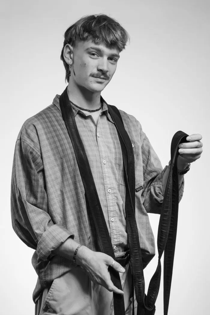 Un jeune homme moustachu, vêtu d'une chemise à carreaux et d'un pantalon clair, porte une ceinture noire autour du cou. Il regarde l'appareil photo avec une expression neutre sur cette photo en noir et blanc.