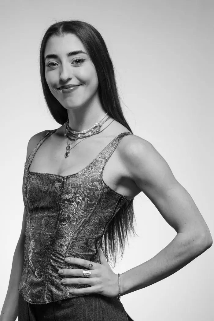 Une jeune femme aux longs cheveux bruns sourit, vêtue d'un haut sans manches à motifs et de colliers superposés. Elle a une main sur la hanche et pose avec assurance sur un fond clair et uni. La photo est en noir et blanc.