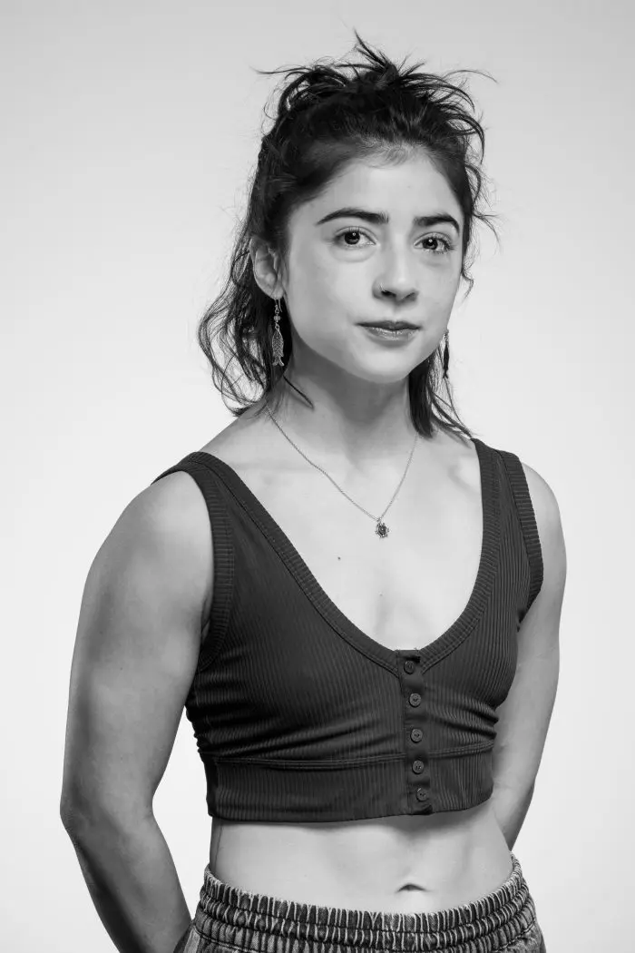 Une jeune femme aux cheveux bruns attachés porte un top sans manches et nervuré ainsi qu'un pantalon à motifs. Elle regarde l'appareil photo avec assurance, sur un fond clair et uni. L'image est en noir et blanc.